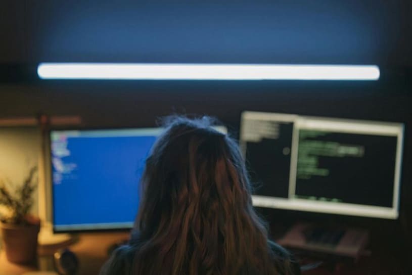Persona trabajando frente a dos monitores con código en pantalla, en un entorno de oficina oscura, ilustrando la labor de un profesional de ciberseguridad dentro del marco europeo de competencias ECSF.